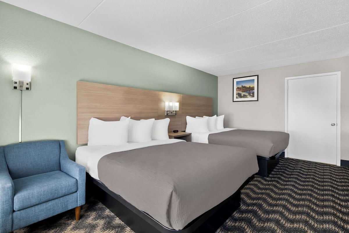 A hotel room with two beds, a blue armchair, a headboard with wall-mounted lamps, and a framed picture on the wall.