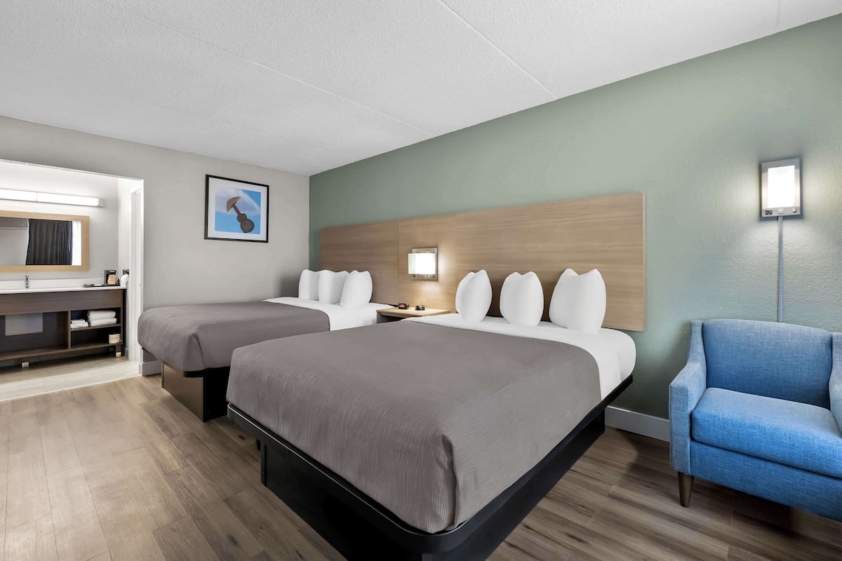 A hotel room with two beds, a desk, a chair, and a wall-mounted lamp.