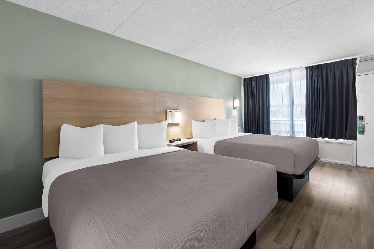 A hotel room with two beds, a wooden headboard, a nightstand with a lamp, and a window with curtains.