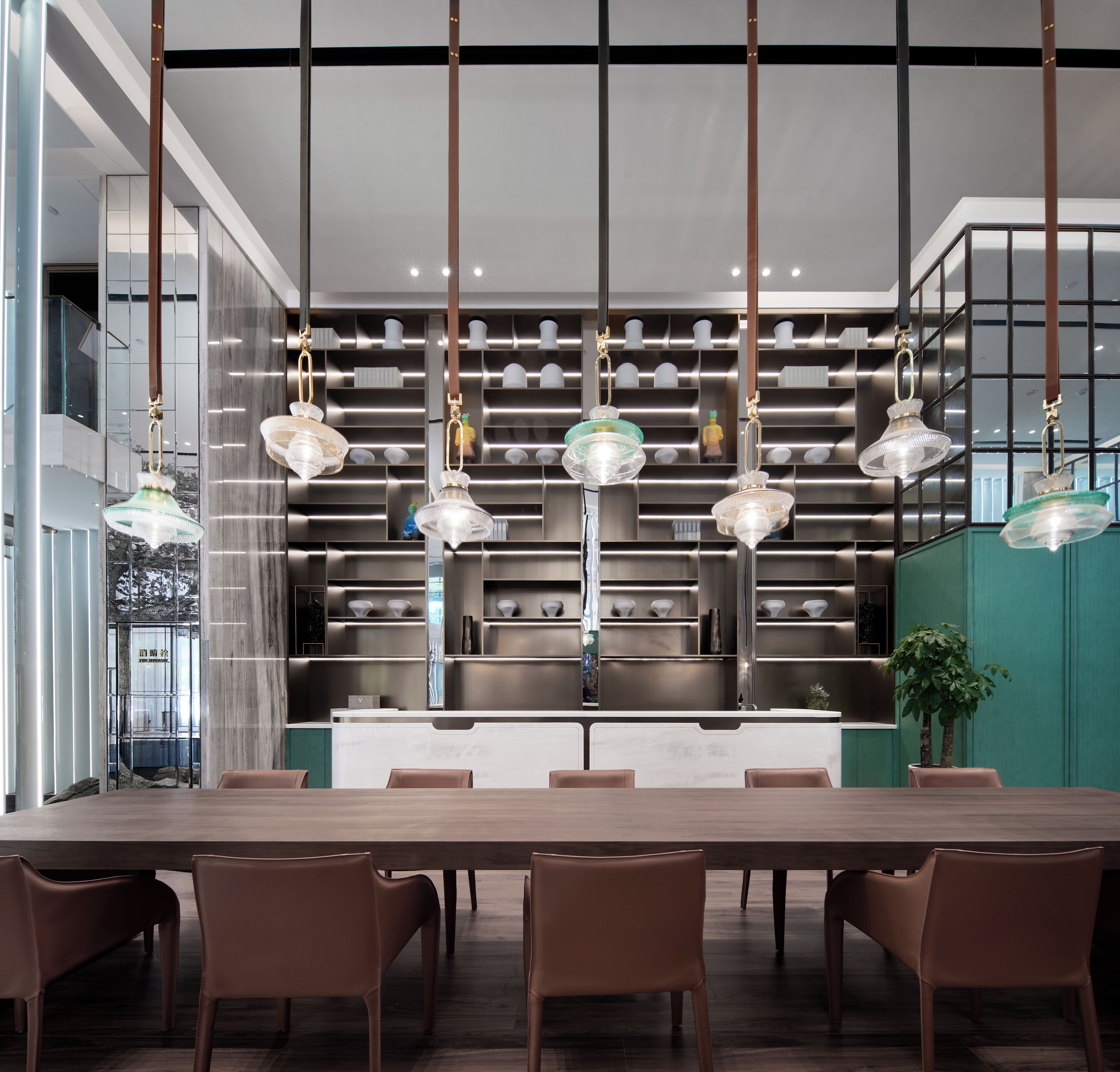 Un espace de repas moderne avec une longue table en bois et des chaises marron, une cuisine équipée d’appareils électroménagers en acier inoxydable et des luminaires suspendus.