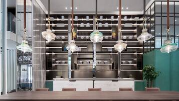 Un espace de repas moderne avec une longue table en bois et des chaises marron, une cuisine équipée d’appareils électroménagers en acier inoxydable et des luminaires suspendus.