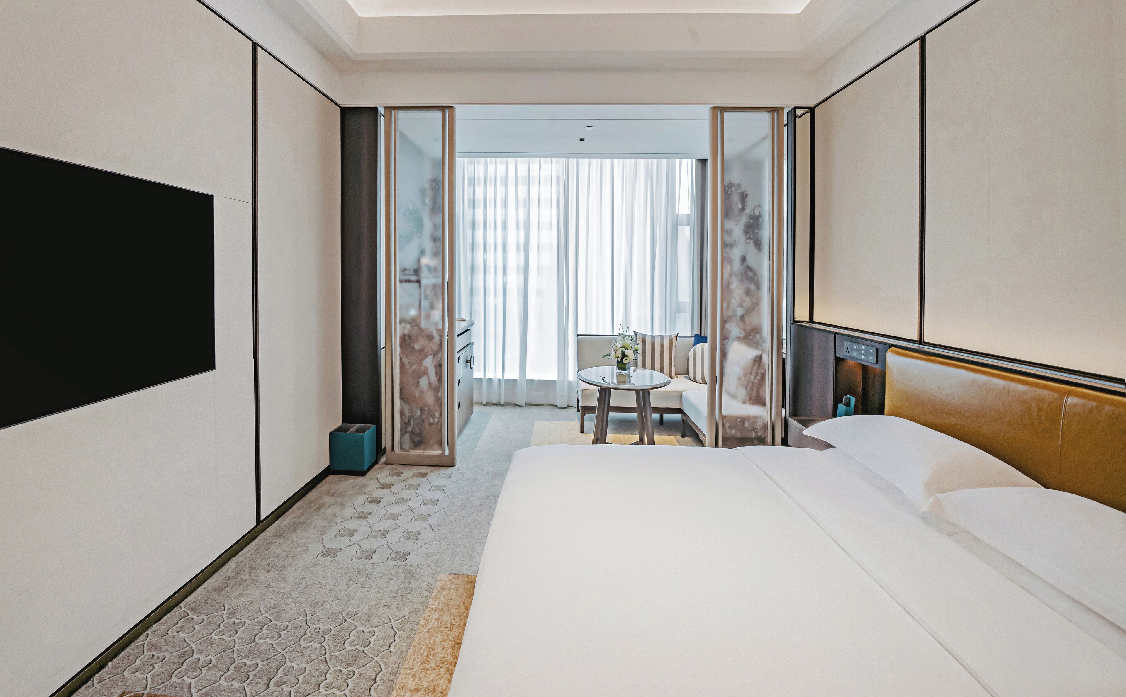 A hotel room with a bed, a round table with a vase of flowers, a seating area with a sofa and curtains, and a large mirror.