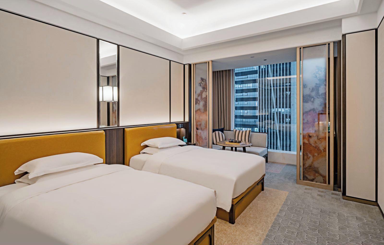 Ein Hotelzimmer mit zwei Betten, einem Schlafsofa, einem kleinen Tisch und einem großen Fenster mit Blick auf ein Gebäude.