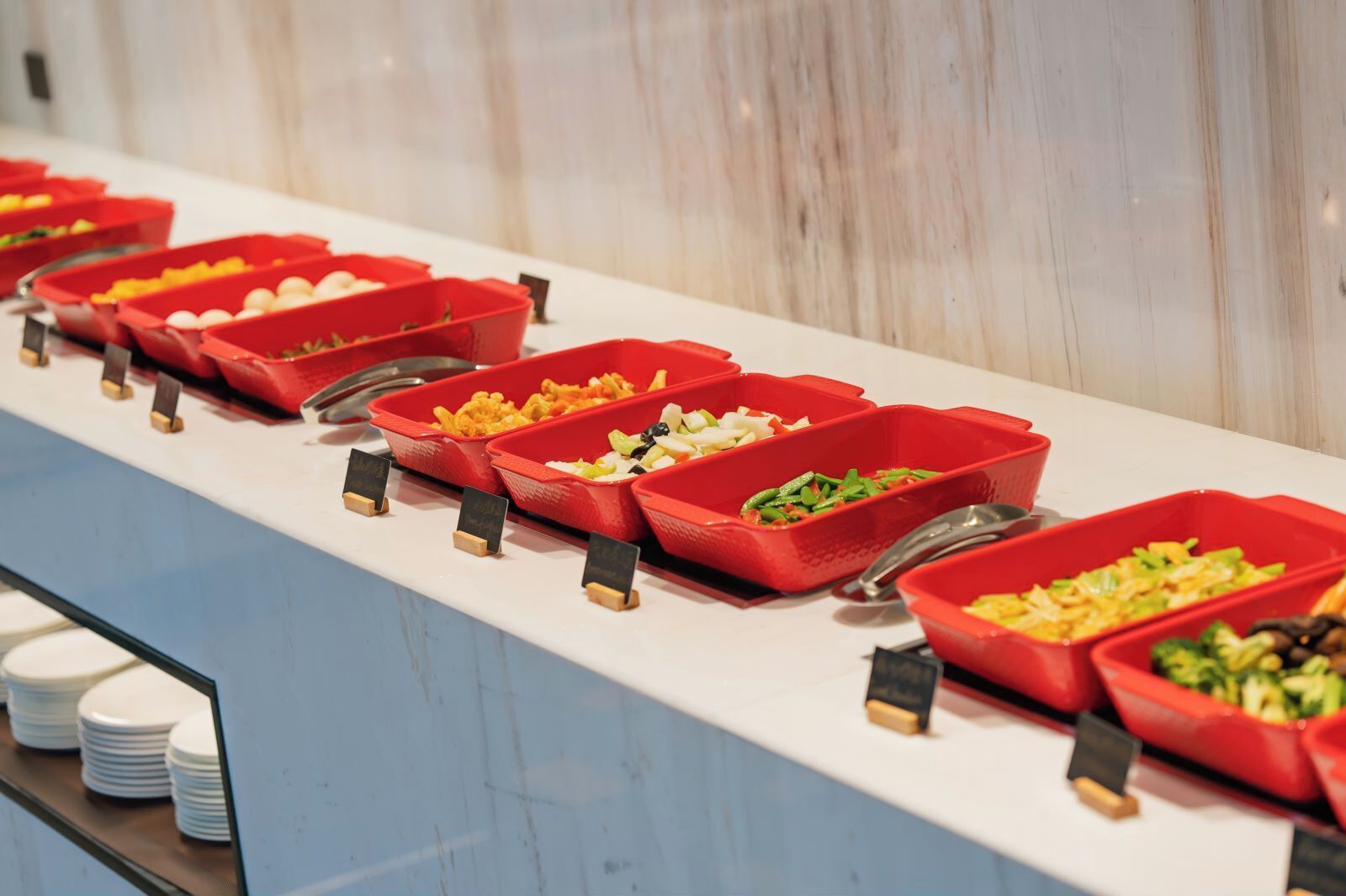 A buffet line with various food items in red trays, arranged on a white counter with a marble wall in the background.