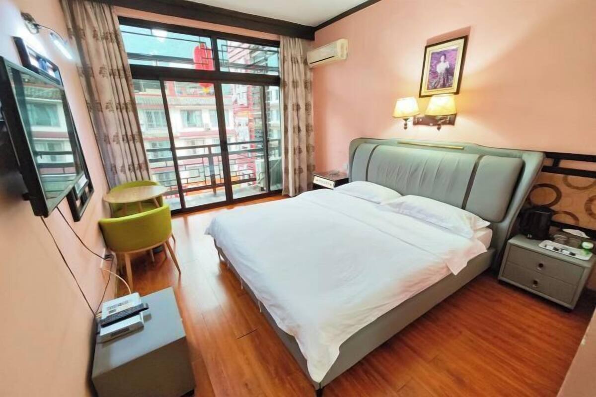 A hotel room with a bed, a desk with a computer, a chair, a nightstand with a lamp, and a window with a view of buildings.