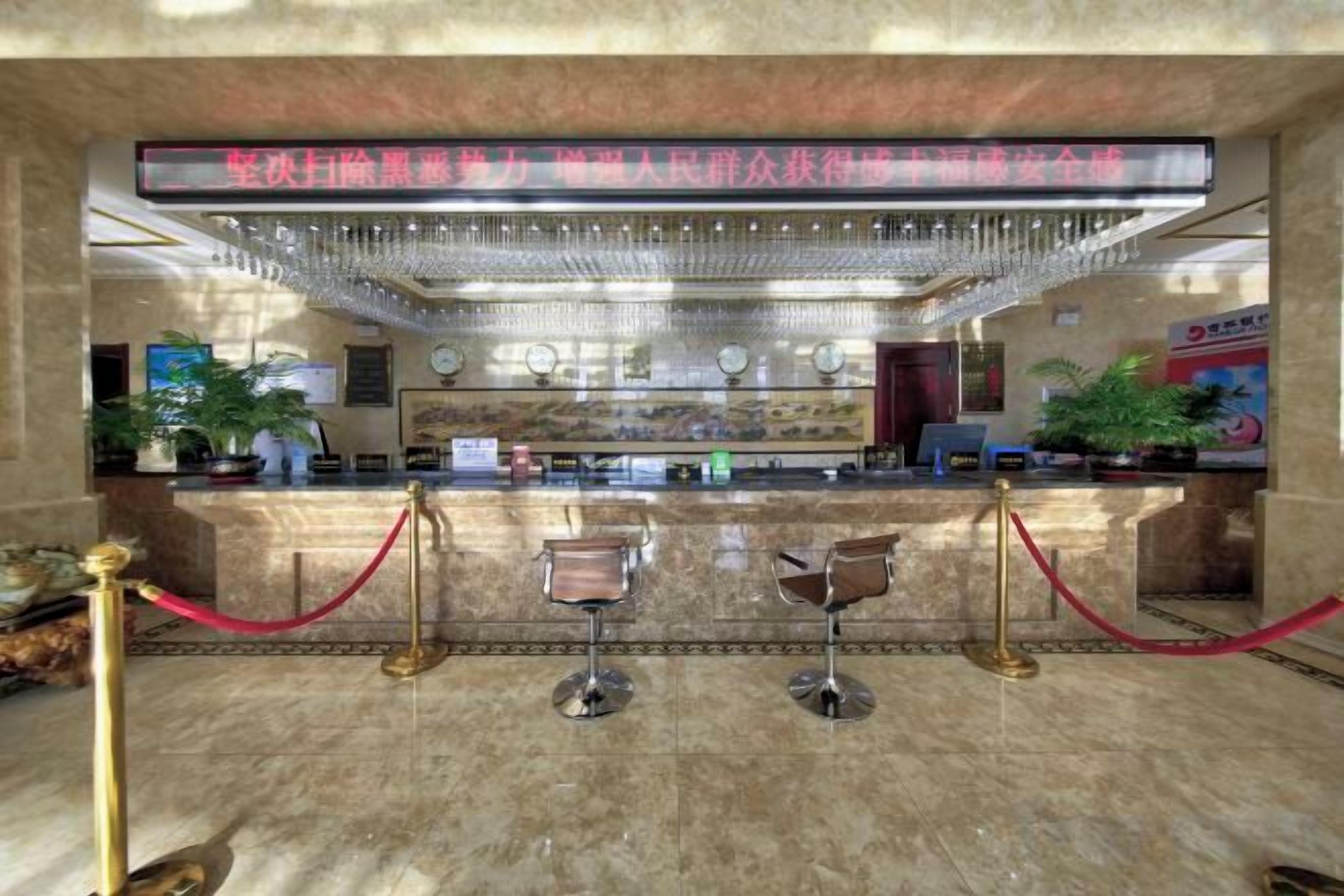 Zona de recepción de un hotel con suelo de mármol, mostrador de recepción, dos taburetes de bar, una barrera de cuerda roja y una araña.