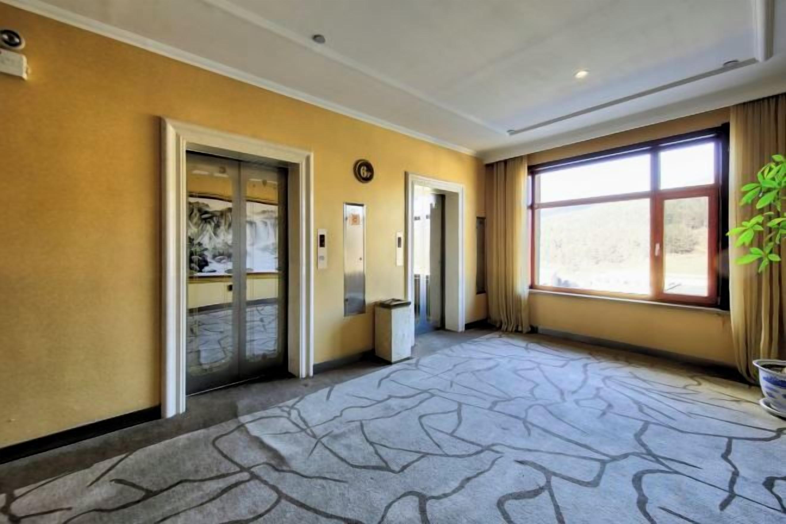 A hotel lobby with a patterned carpet, two elevators, a large window with curtains, and a plant on the right.
