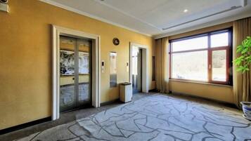A hotel lobby with a patterned carpet, two elevators, a large window with curtains, and a plant on the right.