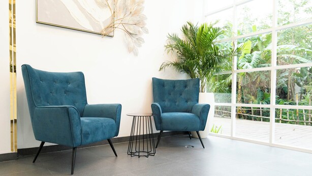 Een kamer met twee blauwe fauteuils, een kleine ronde tafel en een groot raam met uitzicht op groen.