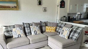 Ein graues Ecksofa mit vielen Kissen in verschiedenen Mustern, eine weiße Ziegelwand, ein kleines gerahmtes Bild und eine Küchenzeile im Hintergrund.