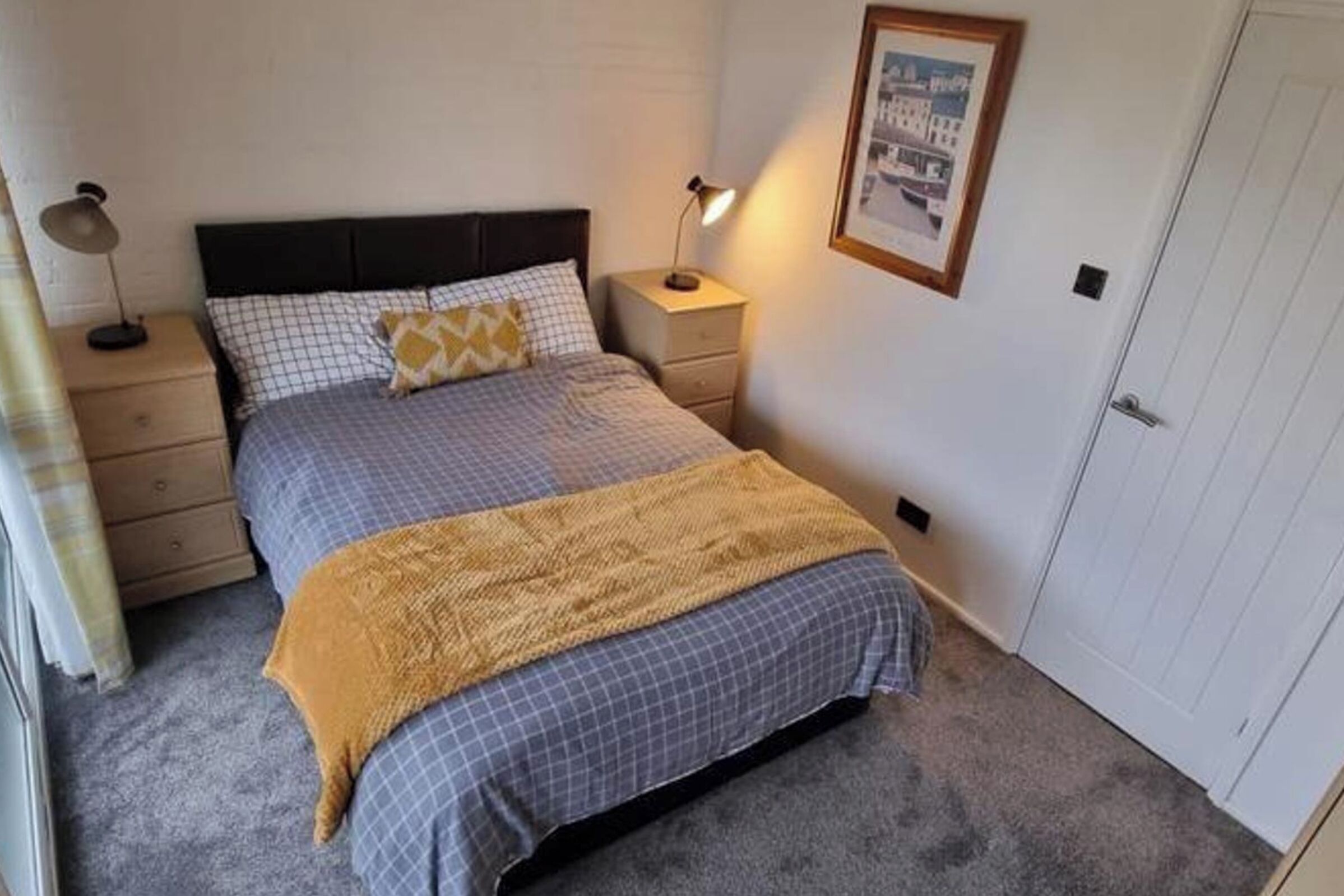A neatly made bed with a checkered duvet, a mustard-colored throw blanket, and two bedside lamps.