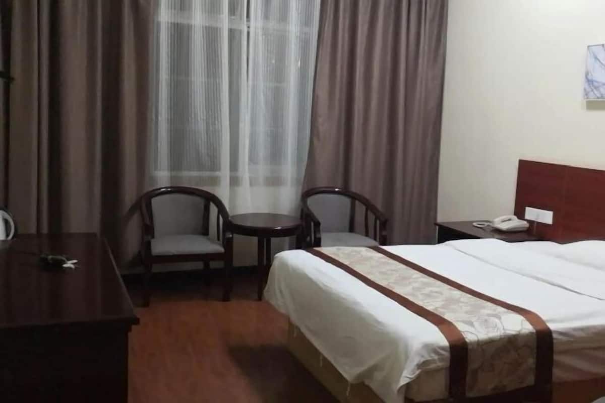 A hotel room with a bed, two chairs, a small table, a nightstand with a phone, and a window with curtains.
