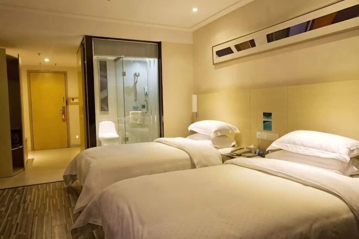 A hotel room with two beds, a glass shower enclosure, a white chair, and a door leading to another room.