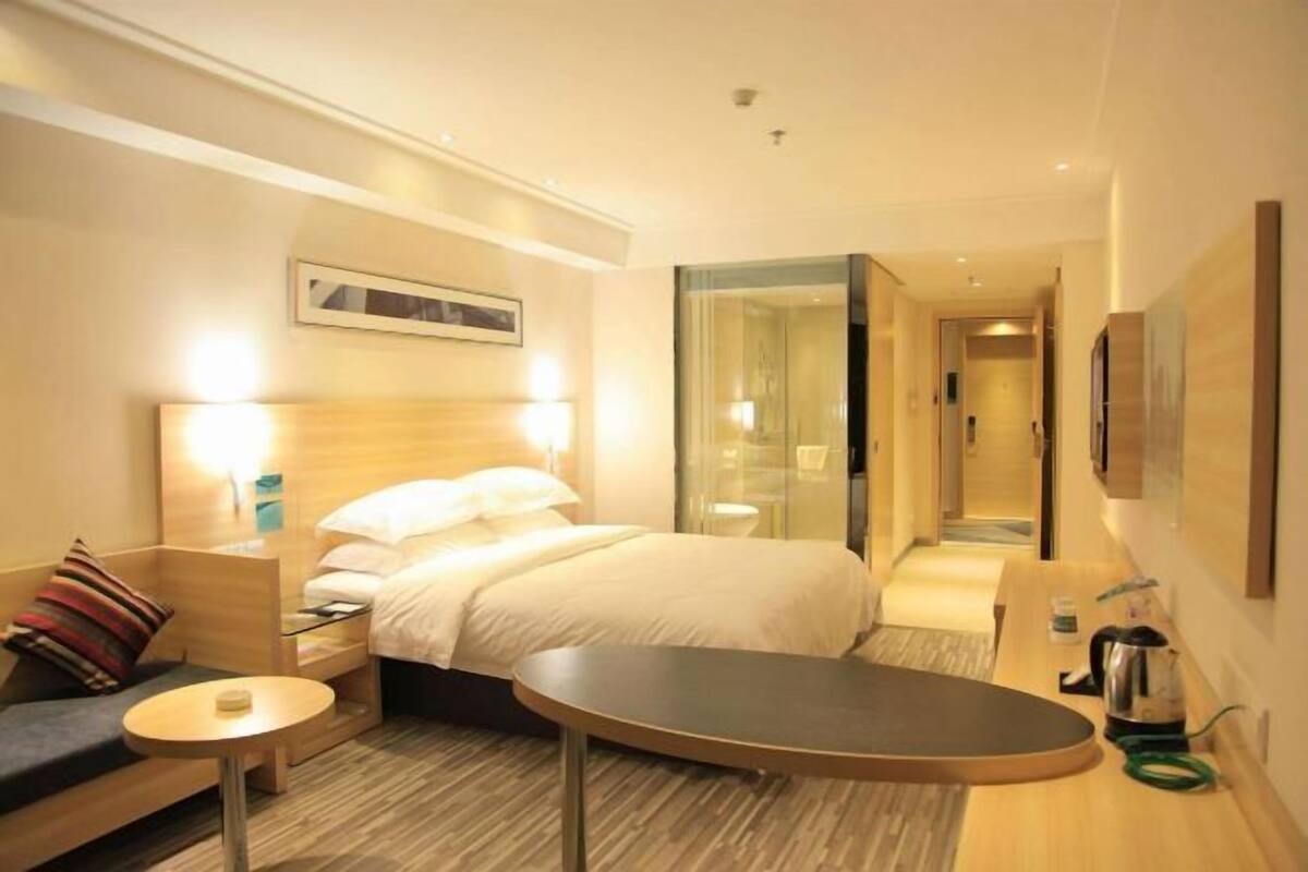 A hotel room with a bed, bedside table, chair, and a small round table. There is a glass partition wall, a door leading to another room, and a wall-mounted TV.