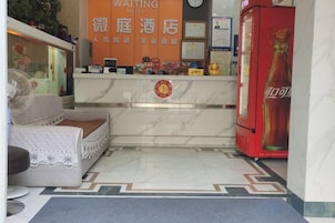 Un área de recepción de hotel con mostrador de mármol, un refrigerador rojo de Coca-Cola y un espacio de espera con sofá y ventilador.
