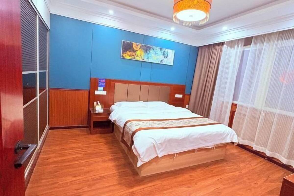 A hotel room with a wooden floor, a bed with a white bedspread, a nightstand, a painting on the wall, and a window with sheer curtains.