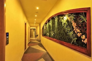 Corredor de hotel com piso coberto por carpete, painel de plantas na parede e janela com moldura de madeira.