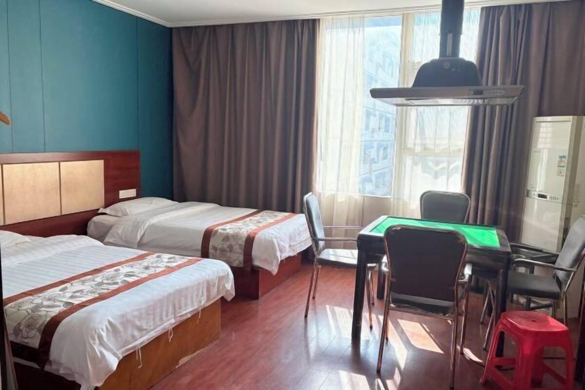 A hotel room with two beds, a desk, chairs, a red stool, and a window with curtains.