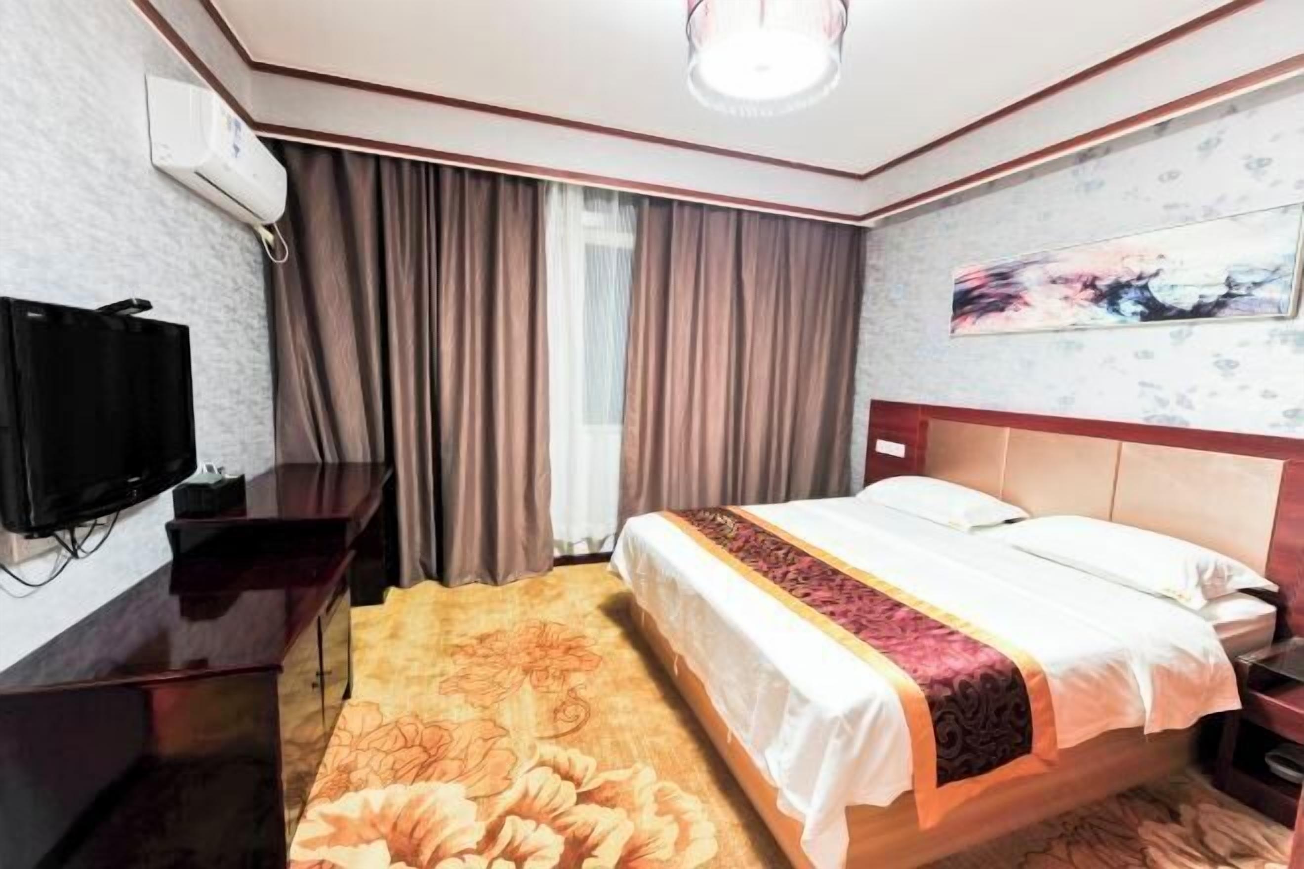 Ein Hotelzimmer mit Bett, Fernseher, Schreibtisch, Stuhl, einem Fenster mit Vorhängen und einem Bild an der Wand.