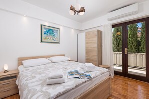 A modern bedroom with a wooden bed, a bedside table with a lamp, a framed painting on the wall, and a view of the outdoors through a glass door. - House Rose With Private Pool And Garden, Pinezići, Croatia (Pinezići)