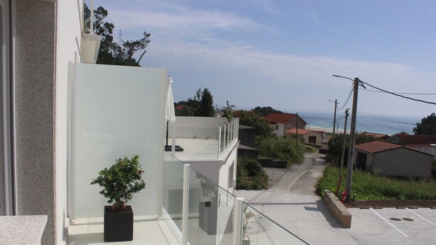 A modern building with a balcony, a potted plant, and a clear view of the ocean and beach in the background.