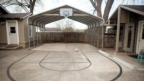 Un terrain de basket en plein air doté d’une zone de sièges abritée, d’une clôture en bois et d’une maison en arrière-plan.