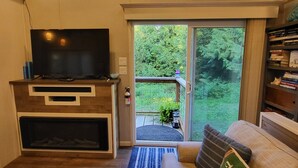 A living room with a flat-screen TV, a fireplace, a sofa with decorative pillows, and a view of a garden through a sliding glass door. - Luxurious quiet cottage on 25 acre rescue farm. In the forest above Port Angeles (Port Angeles)