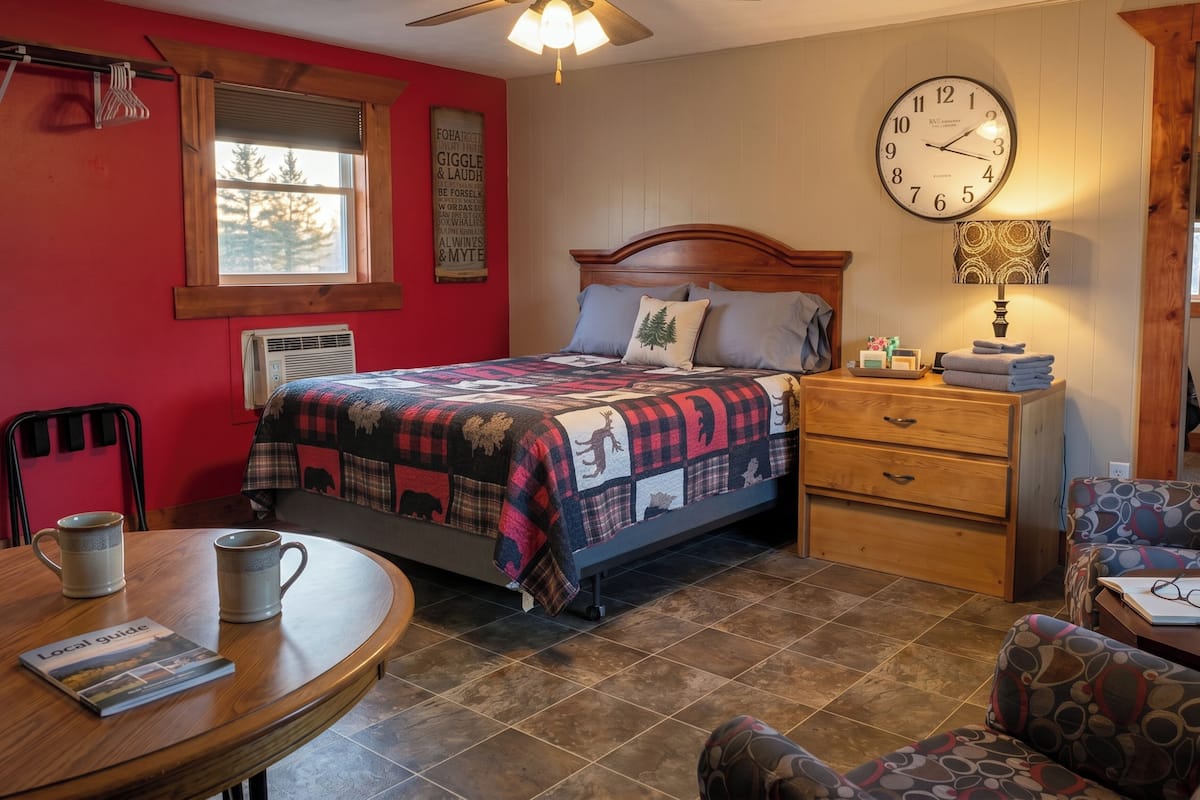 A bedroom with a bed, a wooden nightstand, a round table with two mugs, a clock, and a window with blinds.