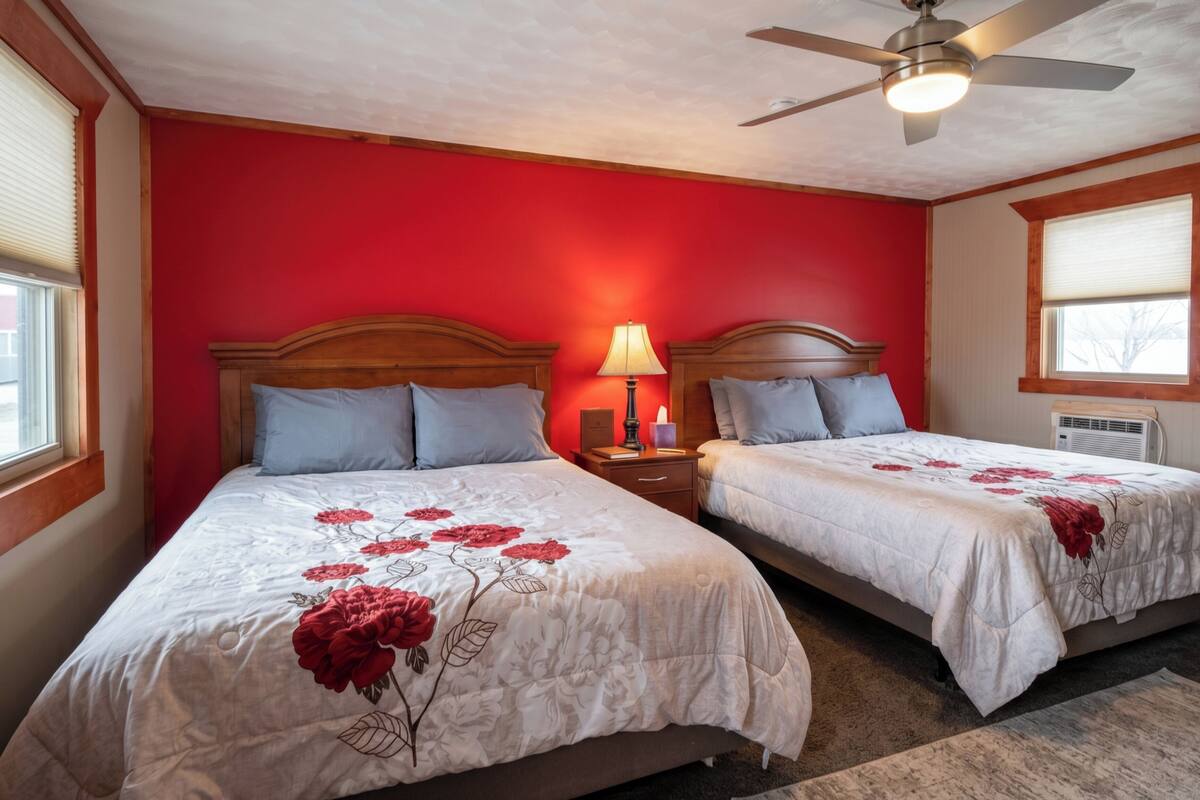 A hotel room with two beds, a ceiling fan, a nightstand with a lamp, and a window with blinds.