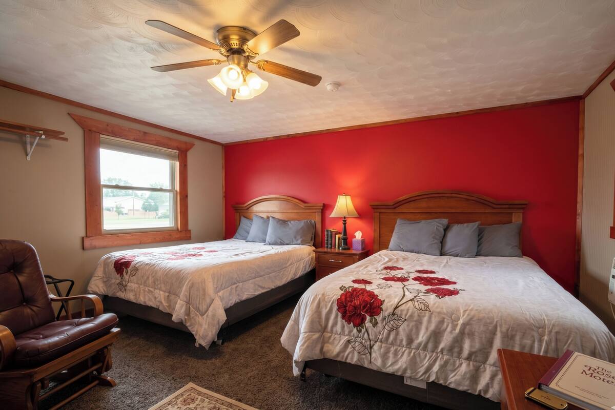A bedroom with two beds, a ceiling fan, a window, a chair, and a bedside table with a lamp.