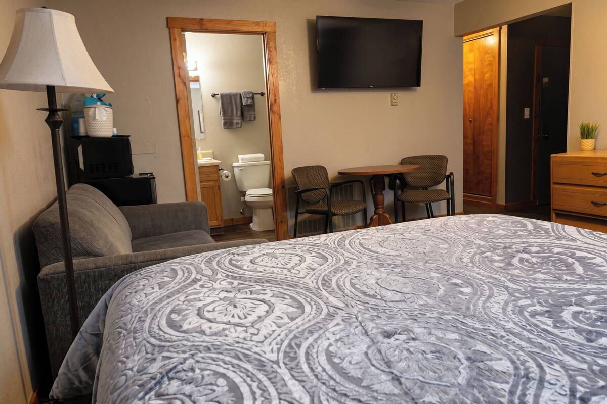 A hotel room with a large bed, a desk, two chairs, a TV, a wardrobe, and a lamp.