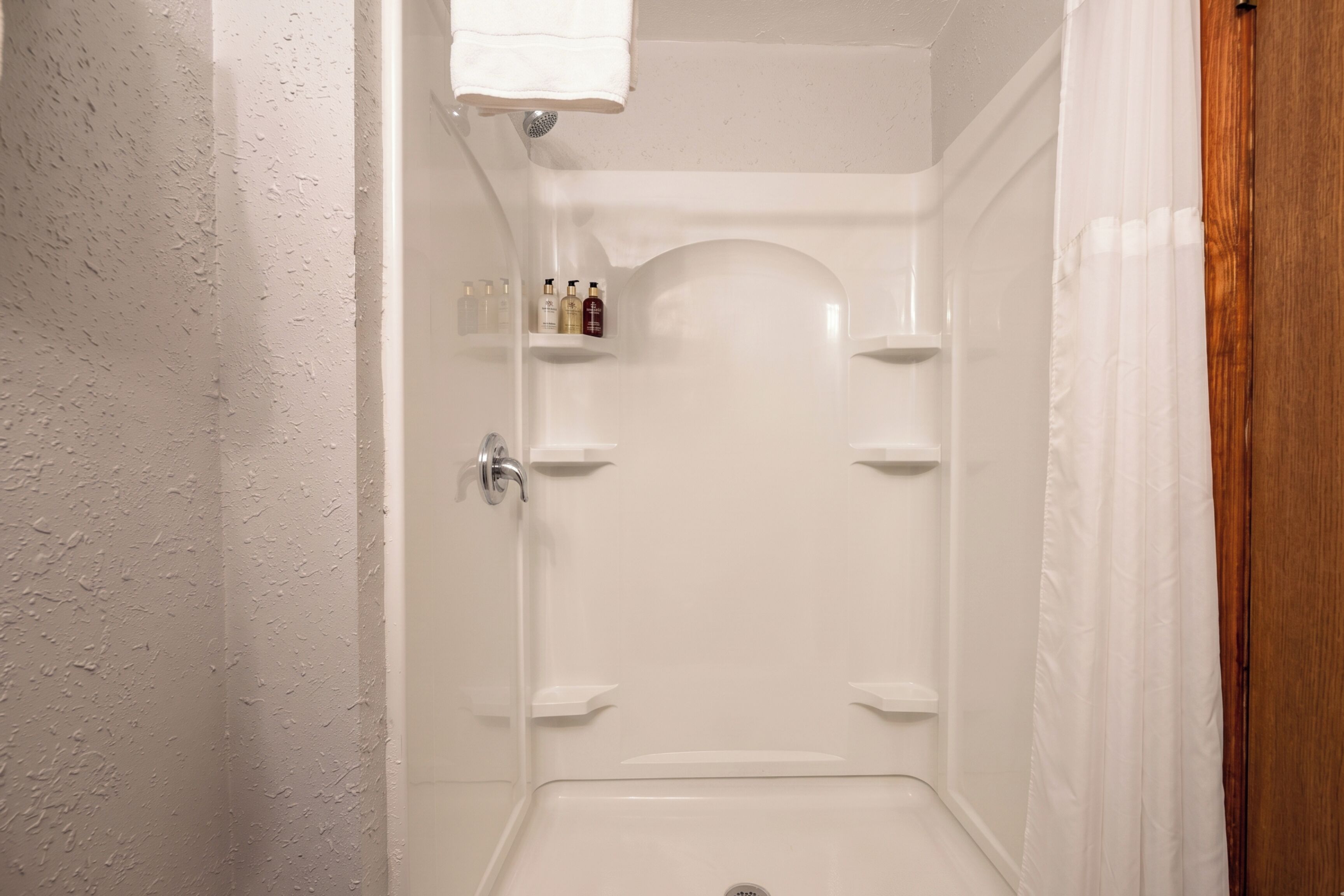 Un box de ducha blanco con cabezal, una toalla colgada en la pared y un estante con artículos de aseo.