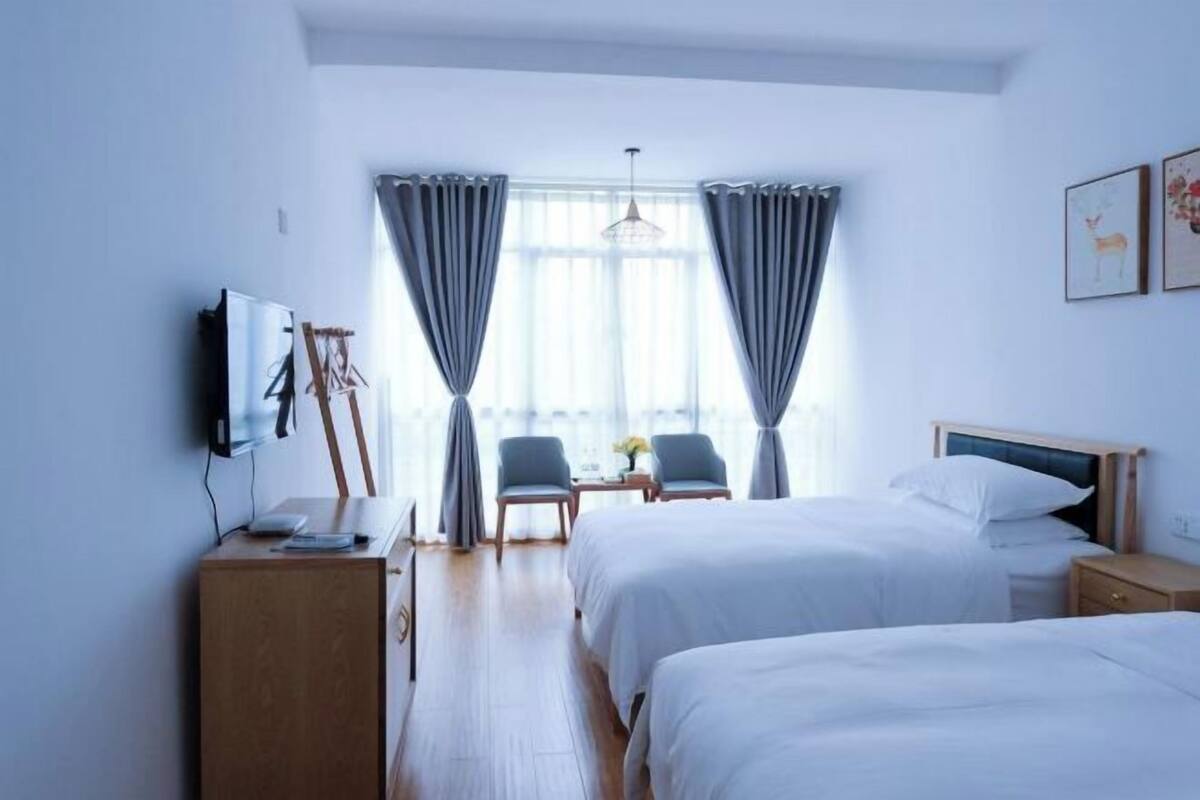 A hotel room with two beds, a desk with chairs, a TV, and a window with curtains.
