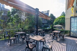 Un patio exterior con mesas y sillas, rodeado de follaje y un edificio al fondo.