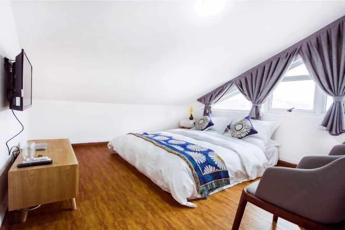 A bedroom with a bed, a wooden nightstand, a chair, a TV mounted on the wall, and a window with curtains.