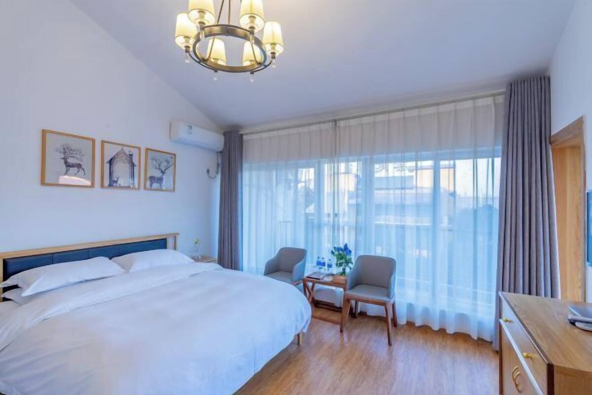 A hotel room with a large bed, a desk, two chairs, a chandelier, and a window with curtains.