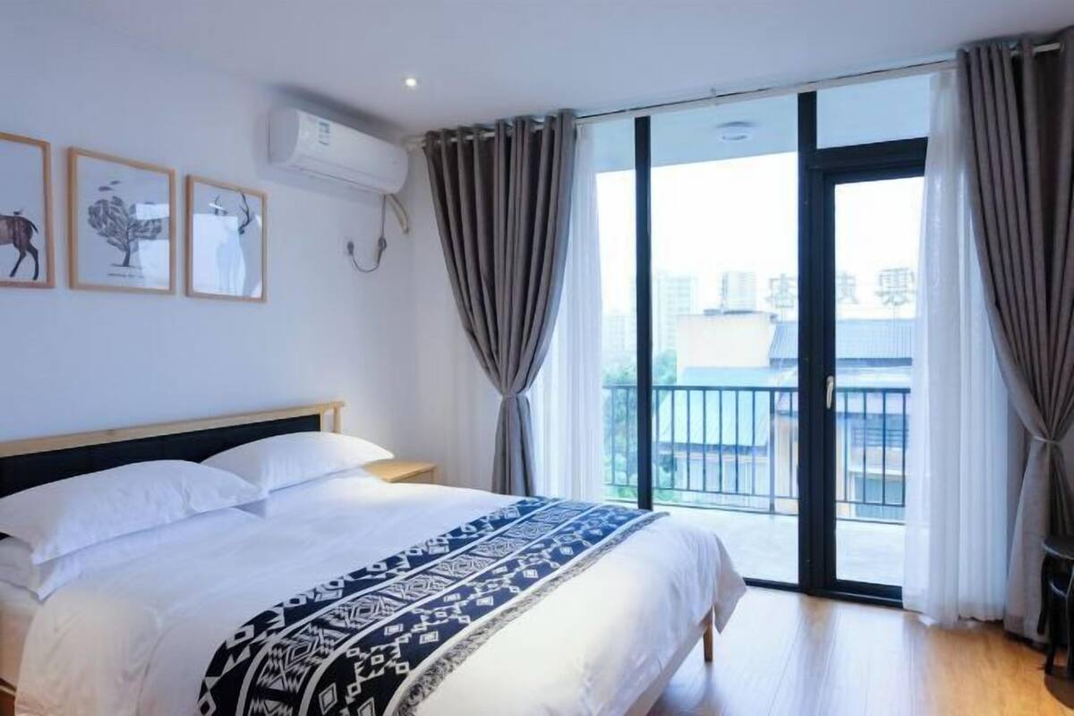 A bedroom with a bed, a window with curtains, a balcony, and framed pictures on the wall.