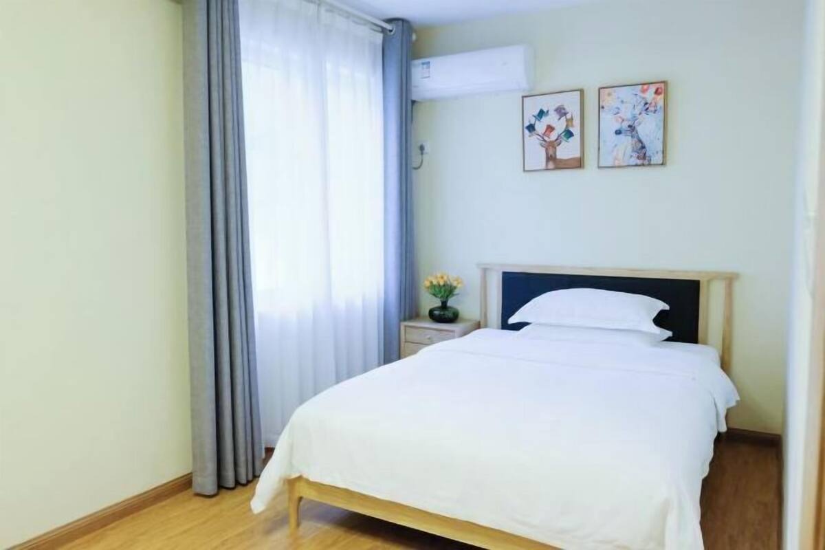 A neatly made bed with a wooden frame and white bedding in a room with light walls, a window with grey curtains, and two framed artworks on the wall.