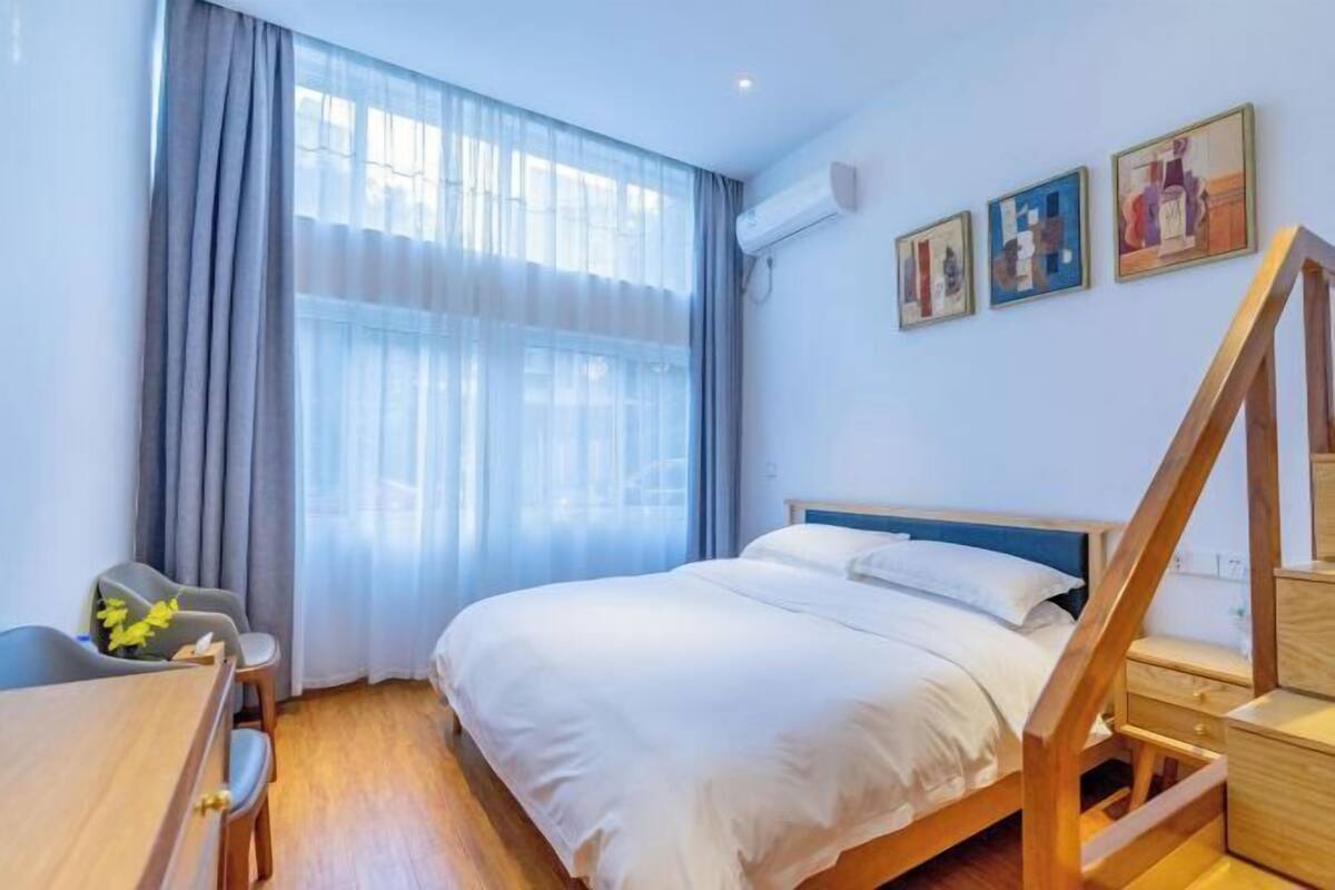 A hotel room with a bed, a desk, and a chair. There are curtains, an air conditioning unit, and artwork on the wall.