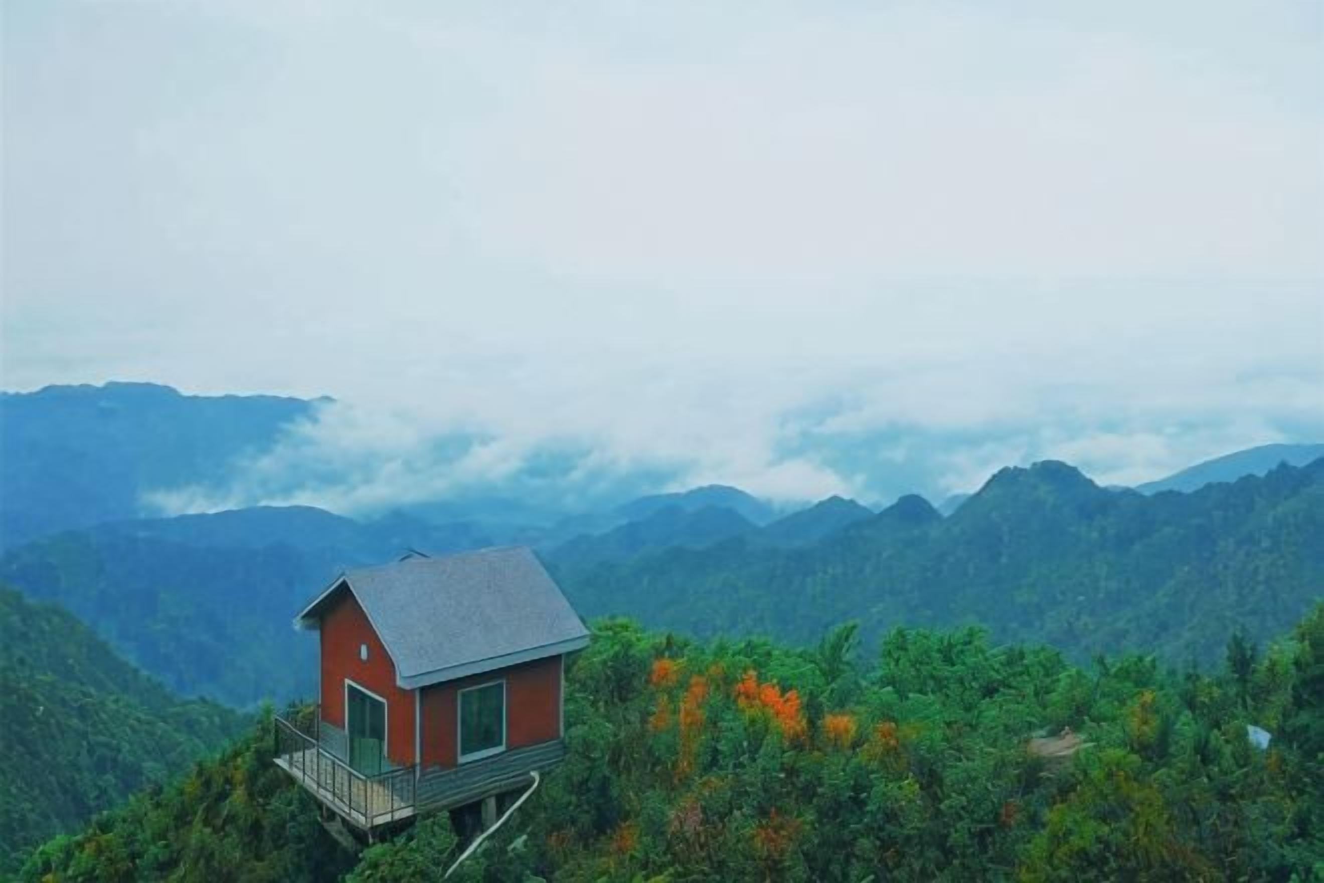 Un pequeño edificio rojo con balcón sobre una montaña, rodeado de un bosque verde denso y montañas distantes.