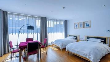 A hotel room with two beds, a dining table with chairs, and large windows with curtains.