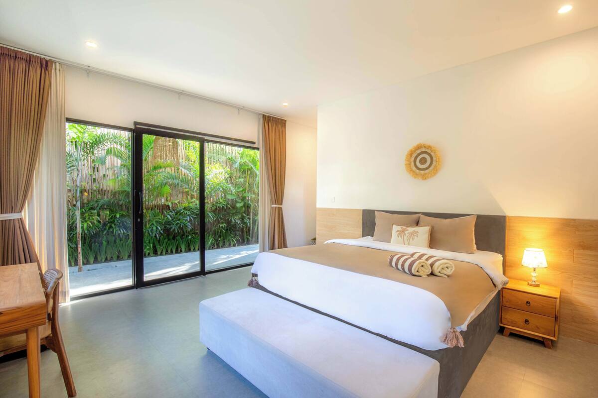 Une chambre spacieuse avec un grand lit, une table de chevet en bois et une vue sur une végétation luxuriante, visible à travers une porte-fenêtre coulissante.