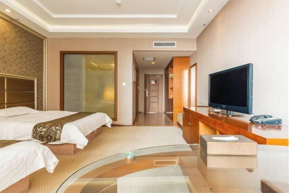A hotel room with two beds, a TV, a desk, and a bathroom visible through a glass partition.