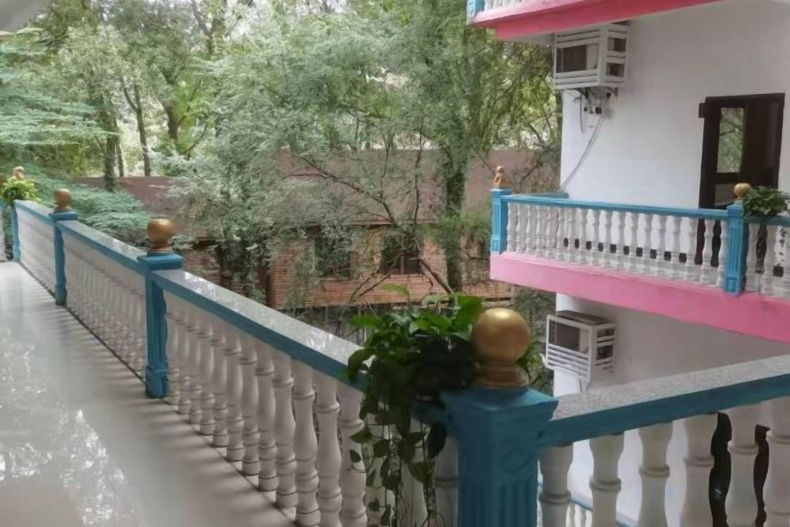 Un balcón con barandilla azul y balaustradas blancas, con vista a un jardín con árboles y un edificio al fondo.