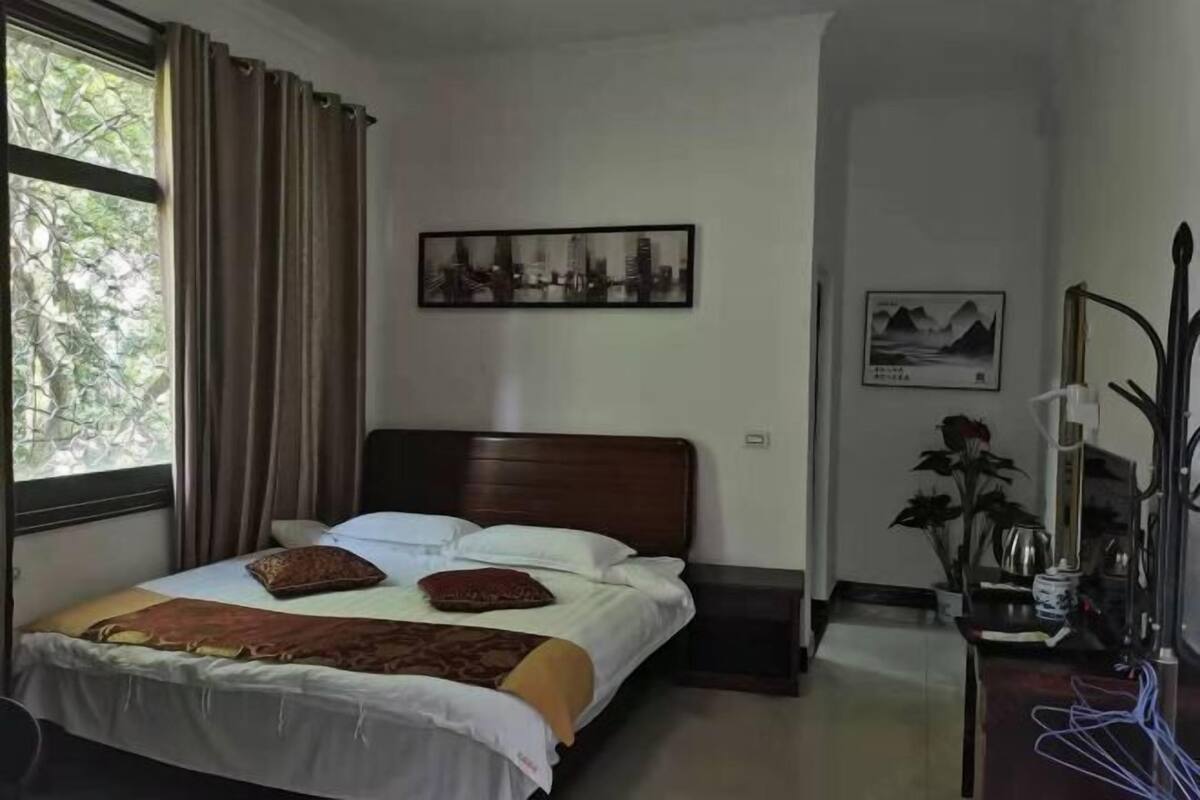 Een hotelkamer met een bed, een raam met gordijnen, een aan de muur hangend schilderij, een nachtkastje met een plant en een bureau met een lamp.