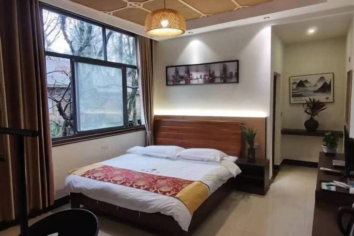 Een slaapkamer met een houten bed, een raam met gordijnen, een kunstwerk aan de muur en een klein bureau met een plantje.