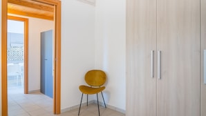Une pièce avec une armoire en bois, une chaise jaune et une porte donnant sur un espace extérieur où se trouvent des chaises blanches.