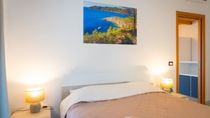 Une chambre à coucher comprenant un lit, deux lampes de chevet, un tableau représentant un paysage côtier accroché au mur, et une porte donnant accès à une salle de bain.