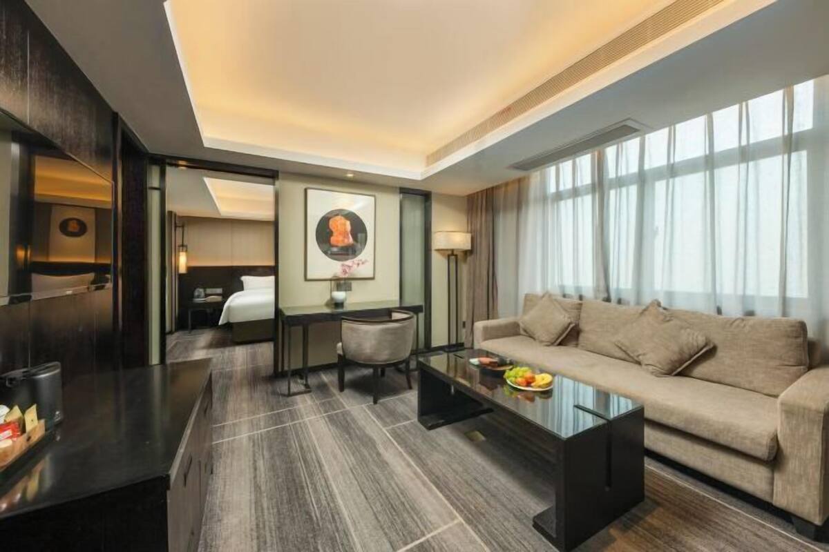A modern hotel room with a sofa, a glass coffee table, a desk, and a bed.