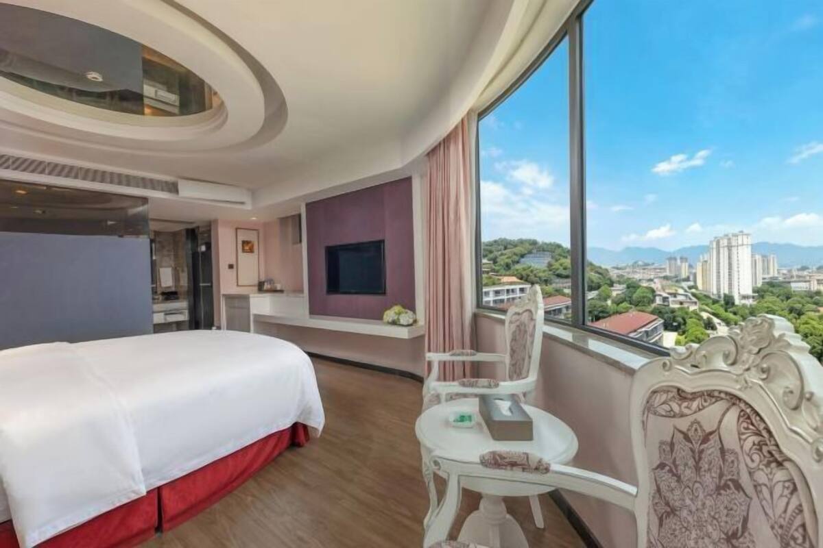 A hotel room with a bed, a small table, a chair, a TV, and a large window with a city view.